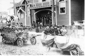 A parade of decorated cars driving by people standing in front of buildings