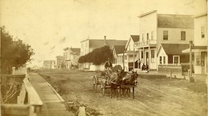 Man in horse-drawn cart on a dirt street lined with buildings on one side