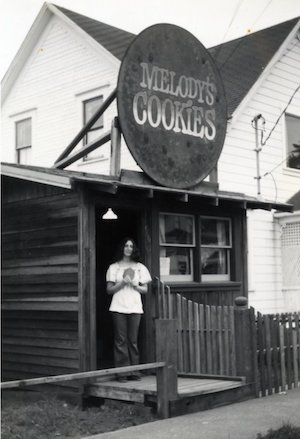 Woman standing in doorway of a building underneath a round sign that says "Melody's Cookies"