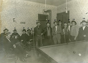 Men standing behind a billiard table, with men seated in the background