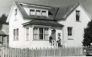 Two people stand outside a two-story house with a wooden fence in front