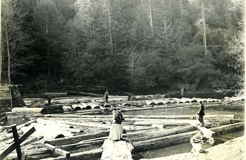 Two women standing on a log. Nearby is a small rowboat and a well dressed man.