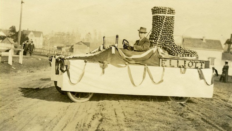 Man driving a parade float, decorated with a large shoe made of apples.