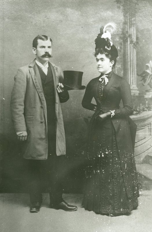 Couple standing together Man has a bushy mustache and is holding a top hat. Woman is wearing a hat with feathers.