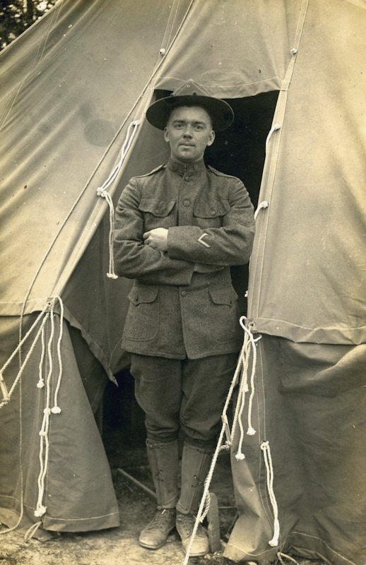 Man dressed in a military uniform standing at entrance of a tent with arms folded across chest