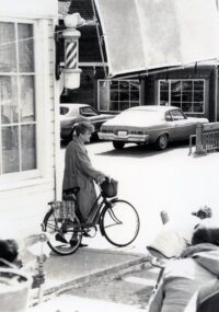 Angela Lansbury stands with a bicycle under a barber pole on a street corner.