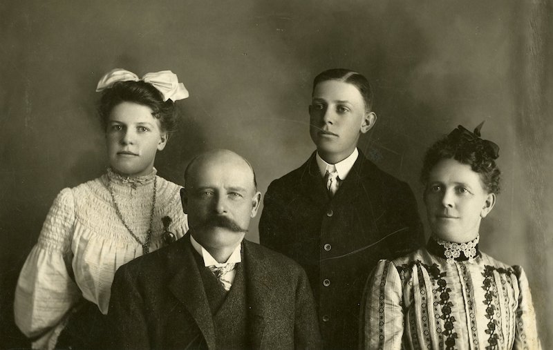 Studio portrait of two adults, a man and woman, sitting with two children, a girl and boy, standing behind them.