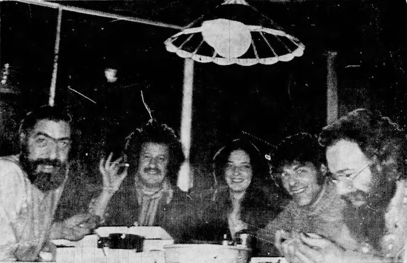 5 people seated at a table, facing camera. One man is holding up a coffee bean for the camera.