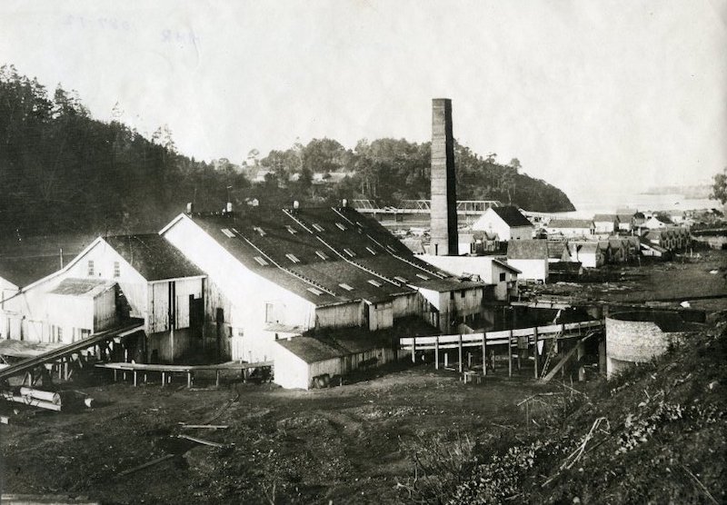 Sawmill sitting on a river flat. In the background, a tall brick chimney and a bridge crossing the river. In the foreground, a large cylinder sits at the end of a conveyor.