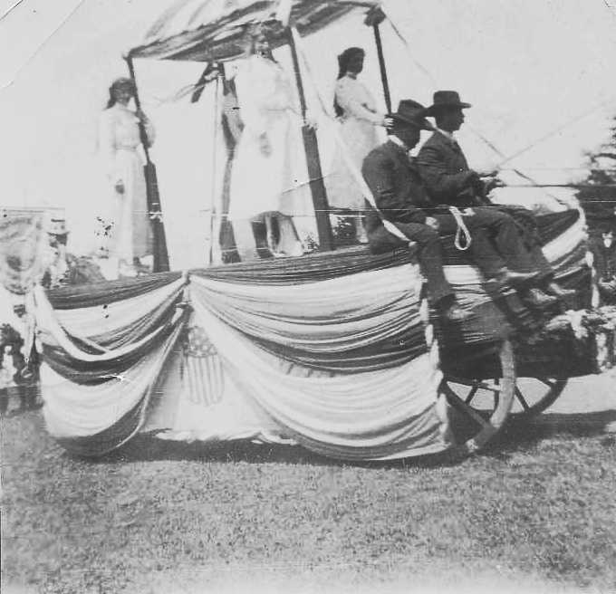 Parade Float with bunting and an American flag. Young girls in white standing on float. Two men are sit up front, driving the wagon.