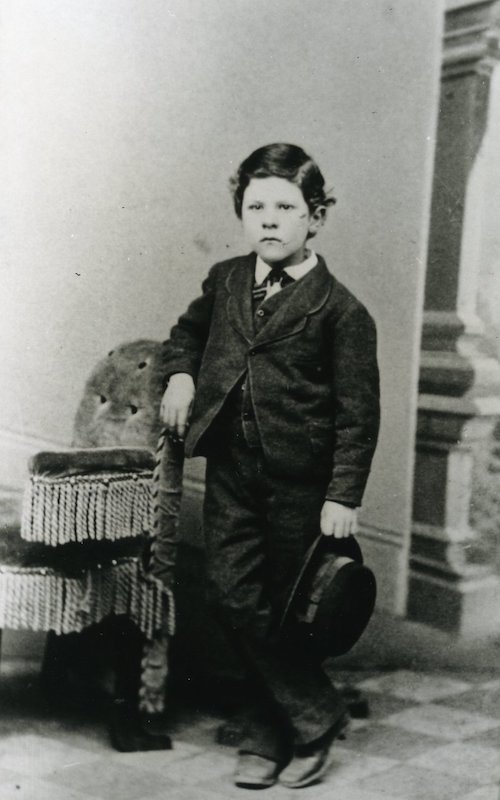Young boy holding hat, standing next to chair