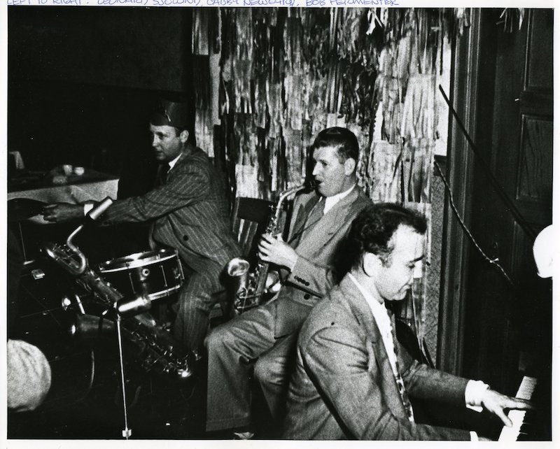Three men seated, playing drums, saxophone, and piano.