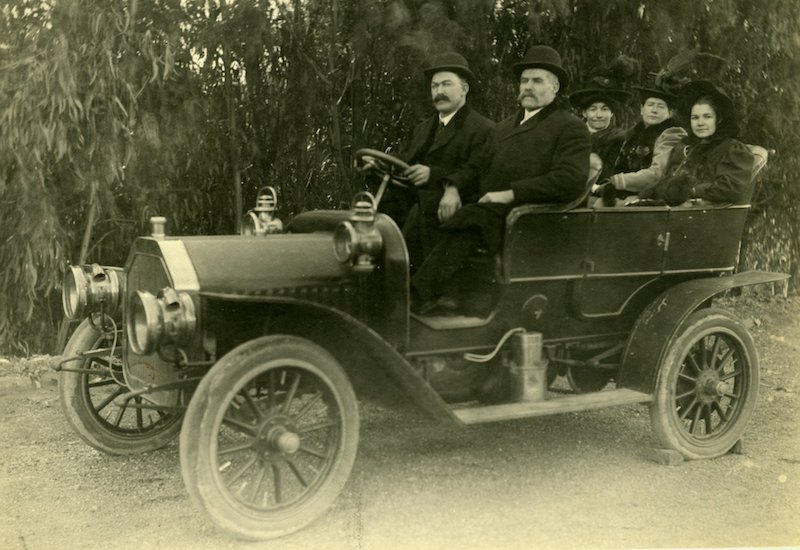 Early-day automobile with two men in front seat and three women in back. Steering wheel is on the right.