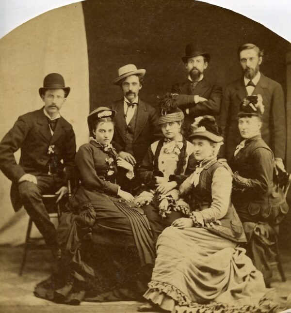 One man seated on a stool and three men standing behind seated ladies, facing camera