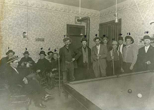 Men holding pool sticks standing behind pool table. A number above each man's head is used to identify him.