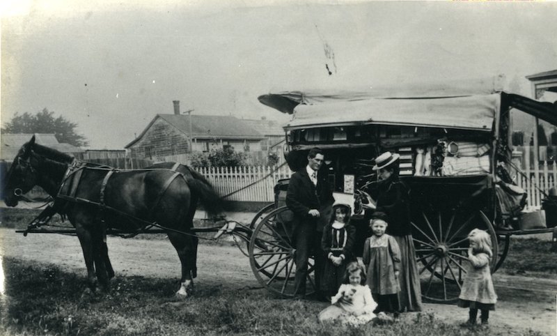 Peddler and customer standing in front of wagon with four children in foreground.