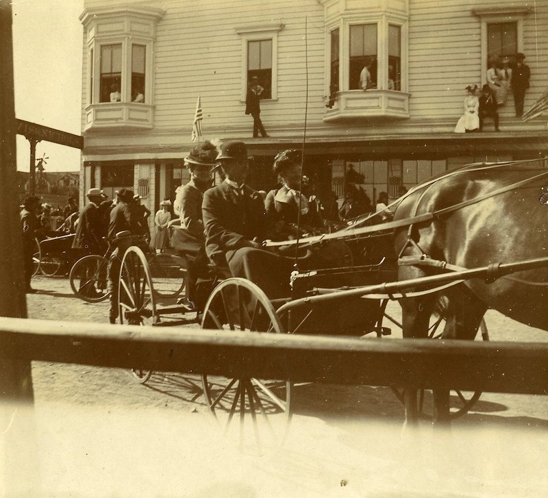 Three people in a horse-drawn carriage. Historic building in background
