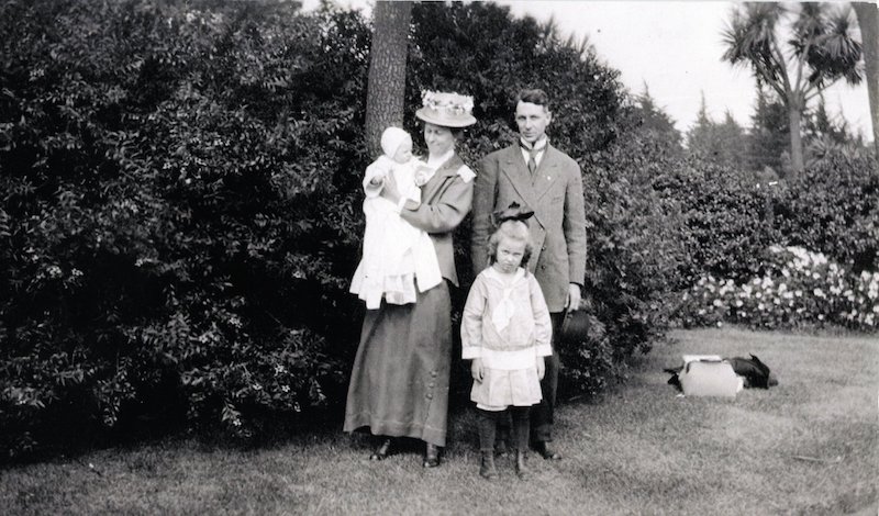 Couple standing in front of tree. Woman is holding a baby and a little girl stands in front of man