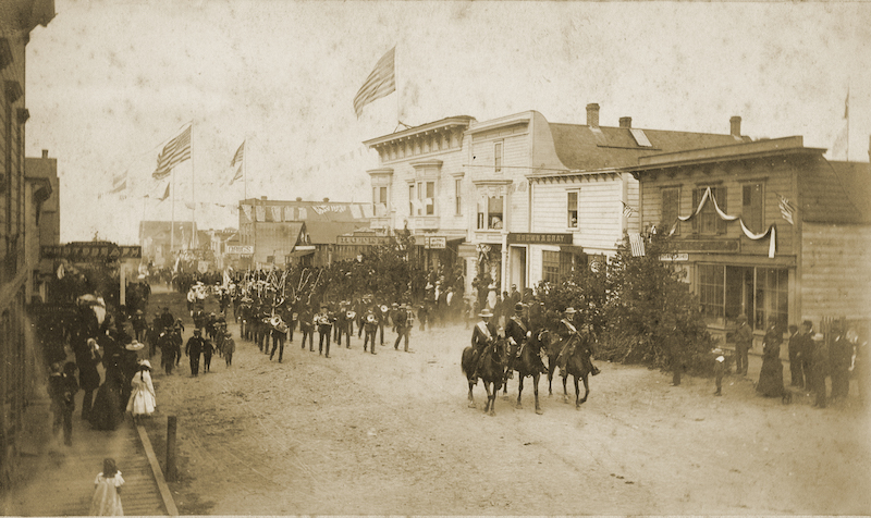 Uniformed men mounted on horses advance eastward on Main Street, followed by a marching band and then ranks of other walking people. Above the street are many large flags waving in the breeze atop very tall "liberty poles.” Both sides of the street are lined with people standing on boardwalks.