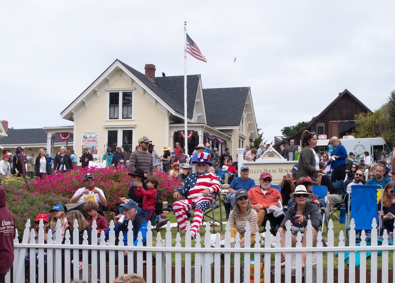 July 4th crowd, decked out in red, white, and blue, sitting on lawn with flag flying in background