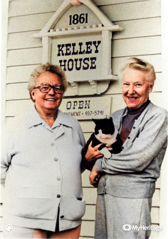 Two women standing in front of a Kelley House sign. One holds a black and white cat.