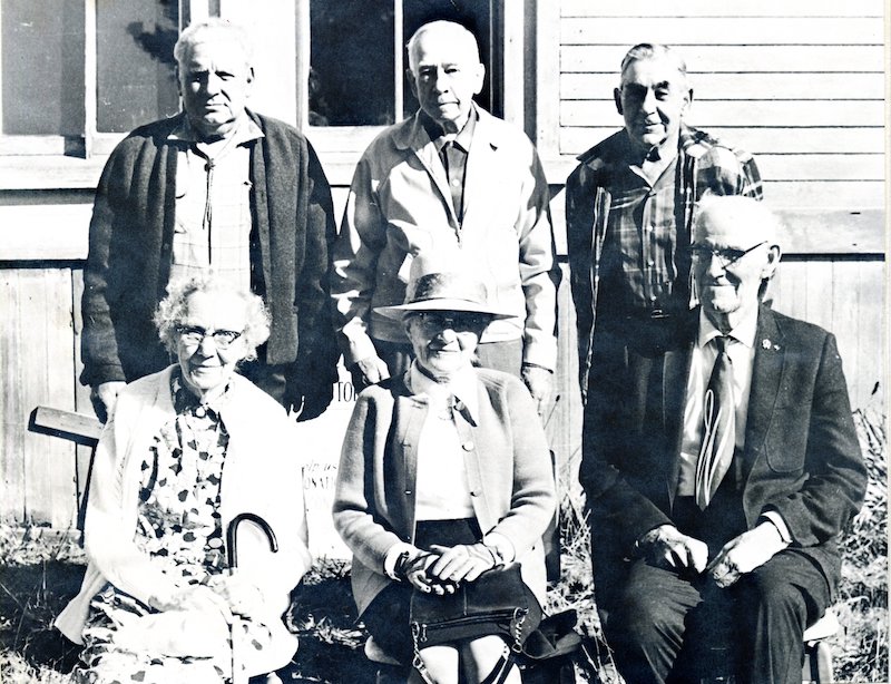 Six people outside a house. 3 men stand behind 2 women and a man who are seated.