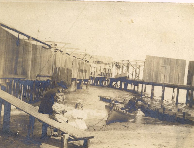 Young girl seated on gangplank, with another child standing, and a man nearby at a pier.