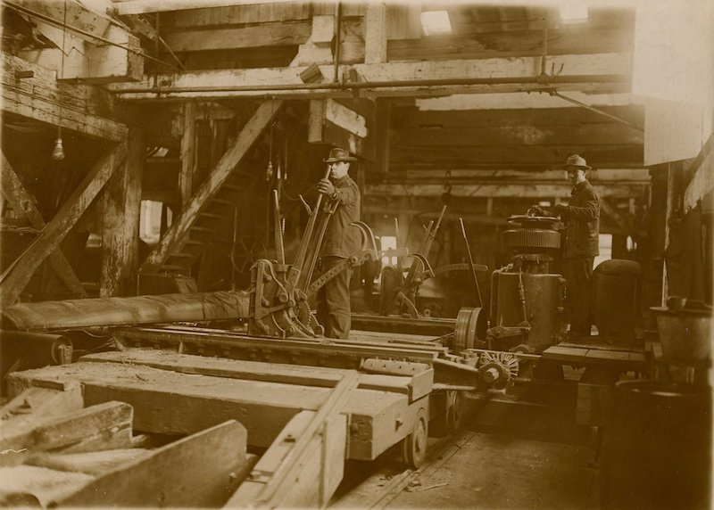 Inside of a saw mill. Two men operate equipment