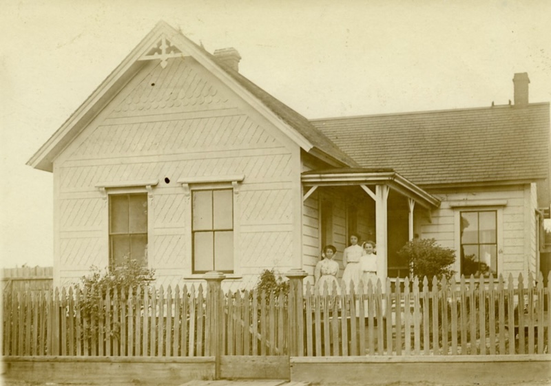 Historic home with picket fence in front. 3 women stand on porch.