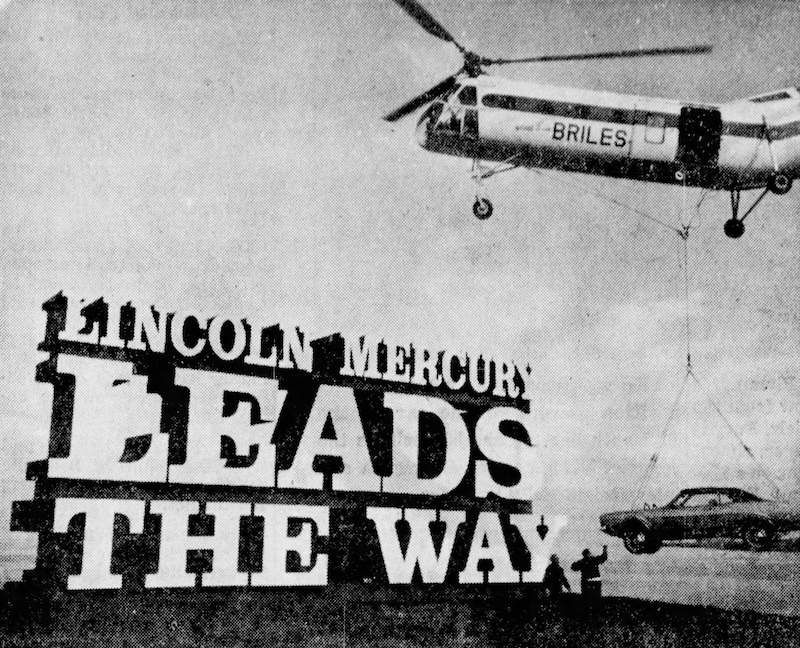 Black and white photo of a helicopter hovering over a large sign that says "Lincoln Mercury Leads the Way." A car is also seen in the photo.