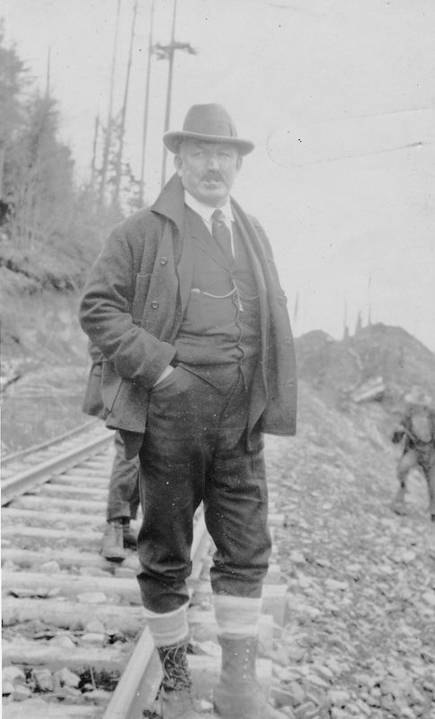 Man standing in a three-piece suit, tie, and boots next to a railroad track