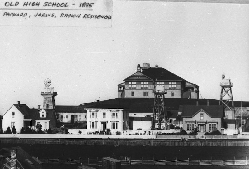 Three historical houses with water towers and windmills. School building on hill in background.