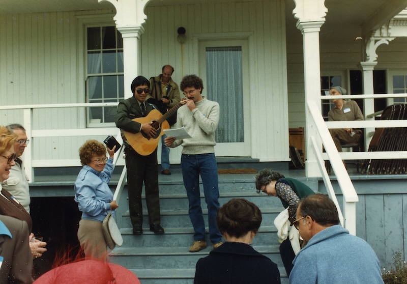 Two men standing on the steps of a historic home. One of them is playing a guitar. A crowd watches them.