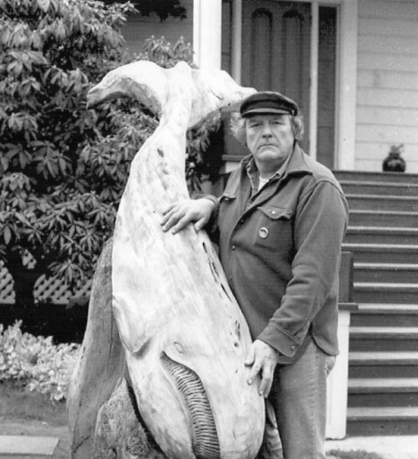 Black and white photo of man in a cap standing next to a sculpture of a whale