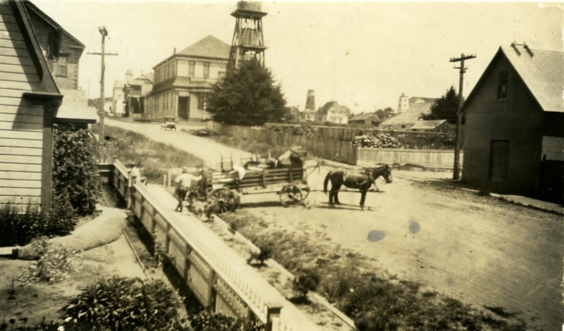 Black and white photo of a historic street with a horse and wagon in the foreground and buildings and a water tower in the background.