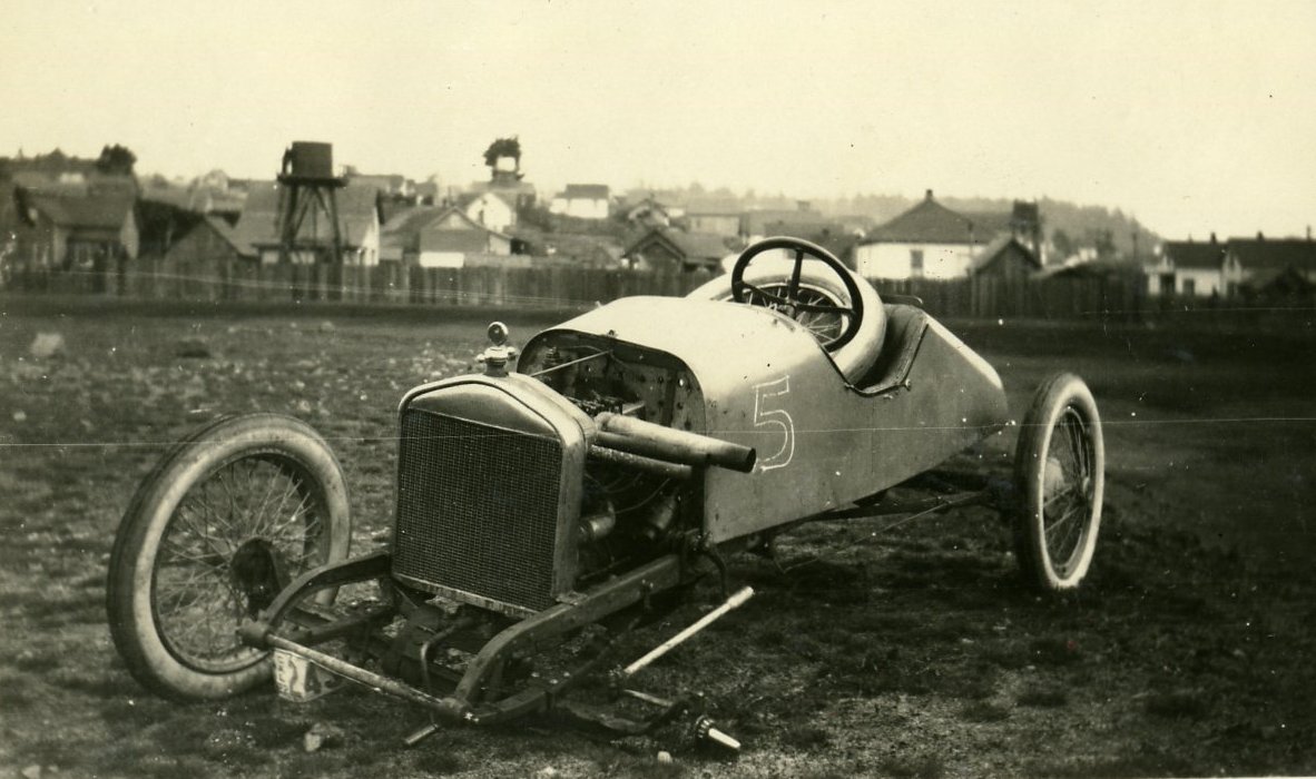 Historical racing car in field, missing a front wheel. Historical buildings in the background.