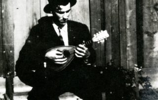 Man seated, wearing jacket and hat, playing a mandolin