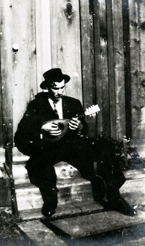 Man seated, wearing jacket and hat, playing a mandolin