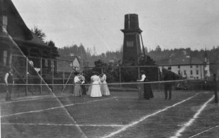 Black and white photograph of men and women in full-length dresses on a tennis court with a water tower in the background