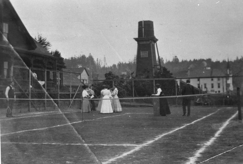 Black and white photograph of men and women in full-length dresses on a tennis court with a water tower in the background