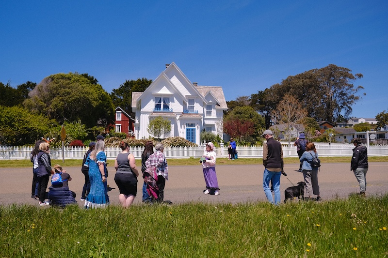 Woman in brightly colored clothes and hat stands in front of historical house with a crowd