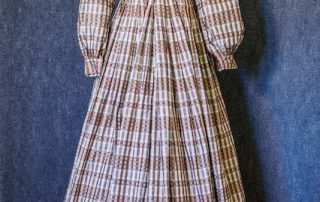 A full-length 19th-century dress displayed on a mannequin. The dress features a plaid pattern, a high white ruffled collar, a fitted bodice with front buttons, long full sleeves gathered at the cuffs, a narrow brown leather belt at the waist, and a floor-length pleated skirt. The garment is photographed against a gray fabric backdrop on a wooden floor.