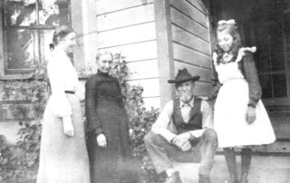 Black-and-white photograph of four people posing on the porch of a wood-sided house. An older man wearing a cowboy hat, vest, and boots sits on the steps beside an older woman in a dark dress. A young woman in a light-colored dress stands at left, while a young girl in a light dress with a large bow stands at right.