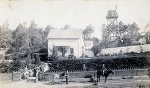 People and horses in front of a house with a fenced-in yard and water tower behind