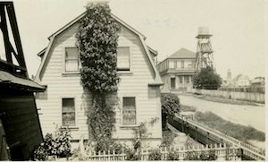 Side view of a two-story house with ivy growing up the side in a small fenced-in yard. A building and water tower are in the background.