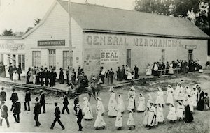 Lines of people in a parade passing a corner building that has "General Merchandise" painted on the side