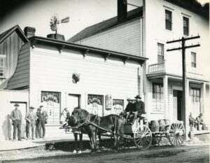 A building with men standing outside and a horse-drawn cart in the street