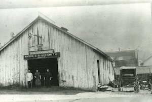 Three men stand in the doorway of a large building with a sign reading "J. A. Chambers Wagon & Blacksmith Shop"