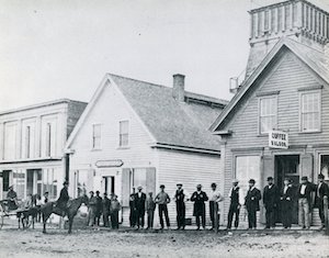 A line of men, with a horse and cart, standing outside a row of buildings  along a street