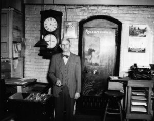 A suited man in a brick room with assorted furniture and a large clock standing in front of a door